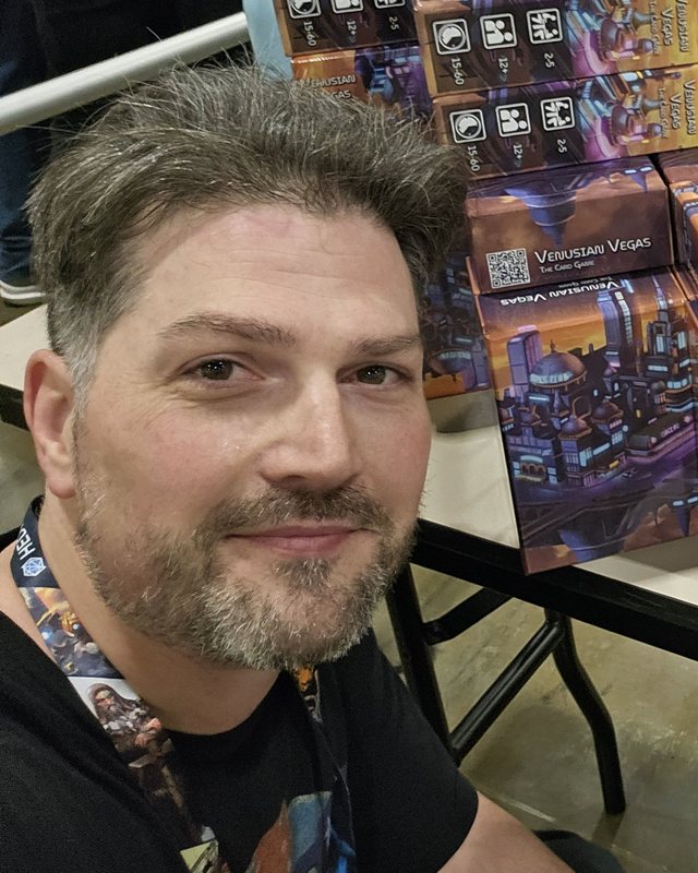 Chris Prime at PAX Unplugged, with stacked boxes of Venusian Vegas behind him.