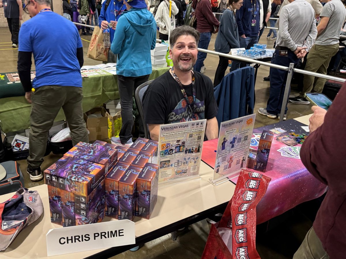 Chris Prime smiling behind his table at PAX Unplugged, with stacks of Venusian Vegas boxes and a Chris Prime name card in front of him.